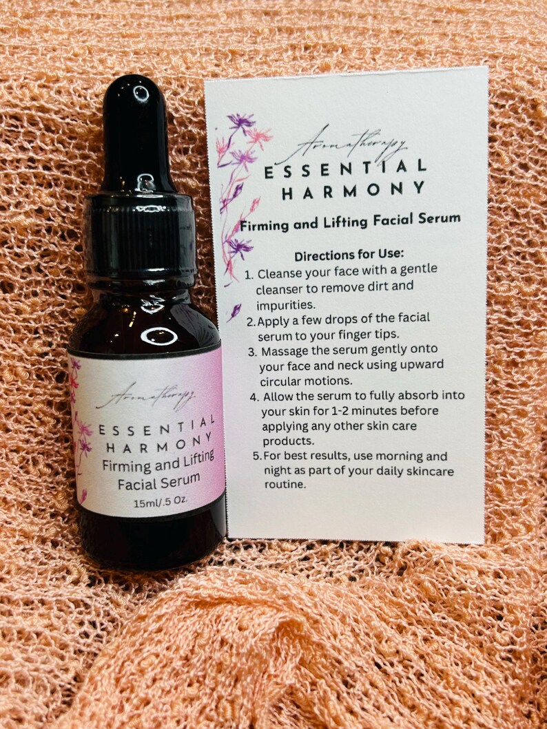 May include: A brown glass bottle with a black dropper and a label that reads "ESSENTIAL HARMONY Firming and Lifting Facial Serum 15ml/.5 Oz." A white card next to it provides usage directions. The background is a peach-colored, textured fabric.
