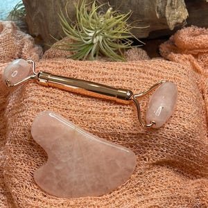 May include: A rose quartz facial roller and a gua sha tool. The roller has a gold-colored handle and two rounded rose quartz stones. The gua sha tool is a smooth, heart-shaped stone. Both are displayed on a peach-colored fabric.