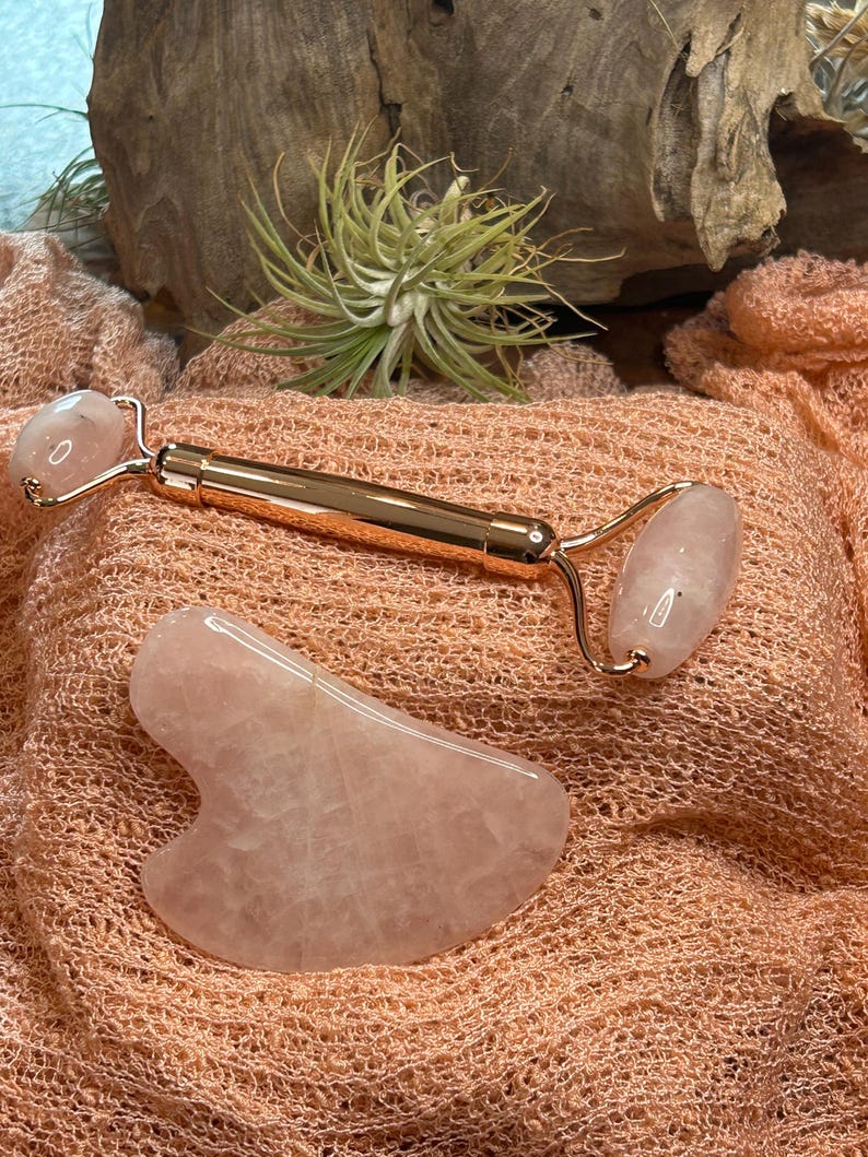 May include: A rose quartz facial roller and a gua sha tool. The roller has a gold-colored handle and two rounded rose quartz stones. The gua sha tool is a smooth, heart-shaped stone. Both are displayed on a peach-colored fabric.