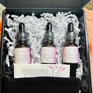 May include: A skincare set in a black box, featuring three brown glass bottles with black droppers. Each bottle has a label with the text "ESSENTIAL HARMONY" and product names. The set includes a small black pouch and a card with instructions.