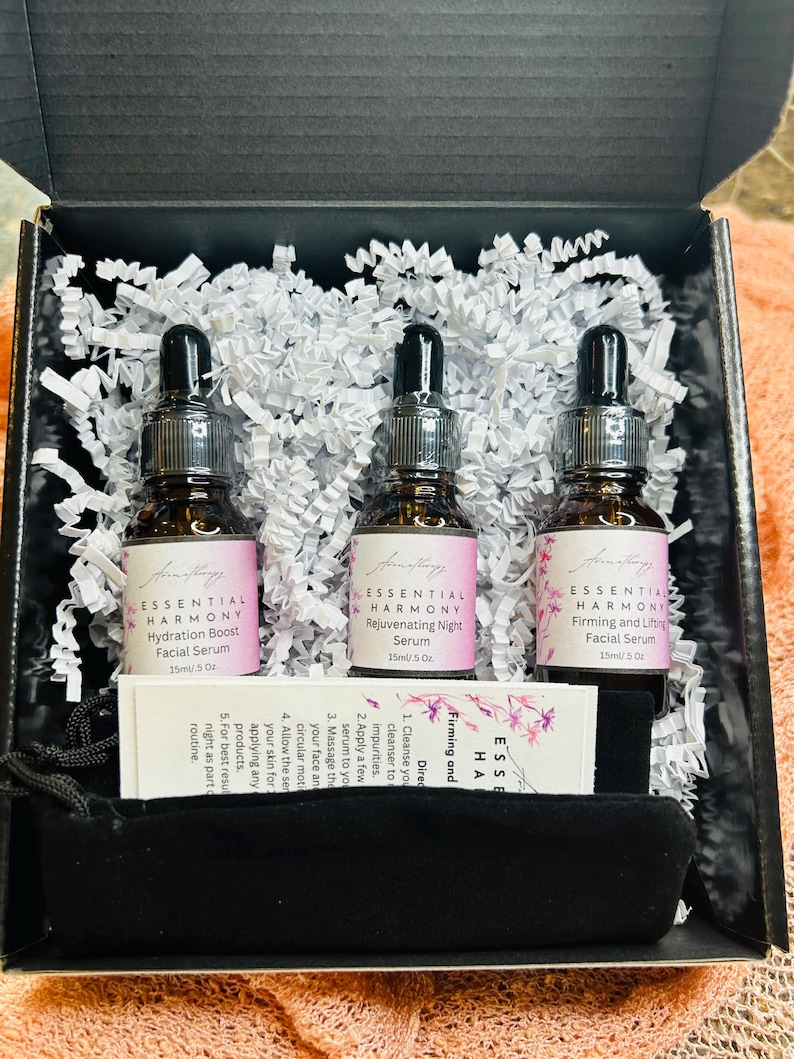 May include: A skincare set in a black box, featuring three brown glass bottles with black droppers. Each bottle has a label with the text "ESSENTIAL HARMONY" and product names. The set includes a small black pouch and a card with instructions.