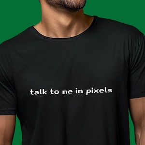 Op de afbeelding: Een zwart T-shirt met de tekst "talk to me in pixels" in wit op de voorkant.