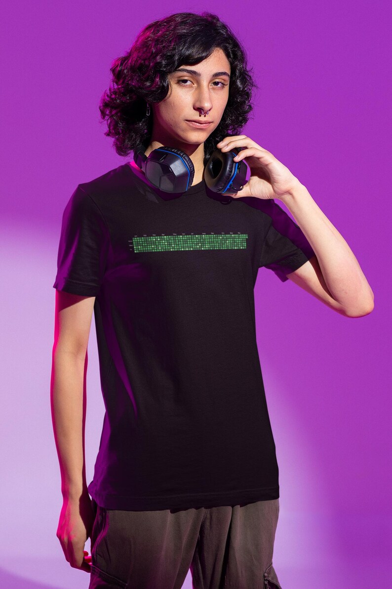 Puede incluir: Una persona con una camiseta negra con un dise&ntilde;o pixelado verde y auriculares azules alrededor del cuello. La persona est&aacute; de pie frente a un fondo morado.