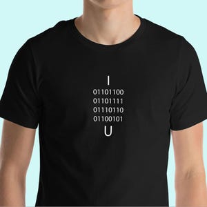 Puede incluir: Una camiseta negra con las palabras "I" y "U" impresas en blanco. Las letras están separadas por una línea vertical de código binario: 01101100, 01101111, 01110110, 01100101.