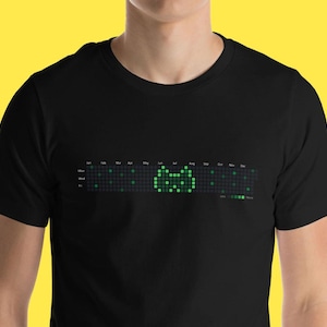Op de afbeelding: Zwart T-shirt met een groene, gepixelde kattenafbeelding. De afbeelding is een kalender met de kat in het midden van de maand mei.