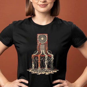 May include: Black t-shirt featuring a graphic design of a stylized, mechanical structure with a fan at the top. The design incorporates red, orange, and cream colors, with dripping chain-like elements. The shirt is short-sleeved.