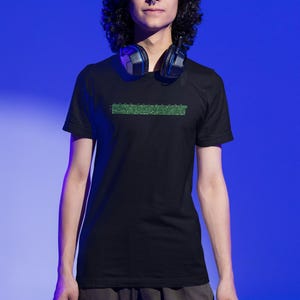 Puede incluir: Una persona con una camiseta negra con una l&iacute;nea de c&oacute;digo verde en la parte delantera. La persona lleva auriculares alrededor del cuello.