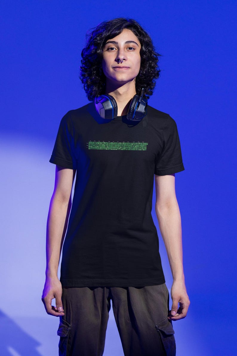 Puede incluir: Una persona con una camiseta negra con una l&iacute;nea de c&oacute;digo verde en la parte delantera. La persona lleva auriculares alrededor del cuello.