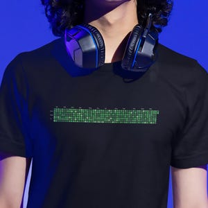 Może przedstawiać: Czarny T-shirt z zielonym pikselowym wzorem z przodu. Wzór przypomina cyfrowy korektor dźwięku lub pasek postępu. Koszulkę nosi osoba z czarnymi włosami i słuchawkami na szyi.