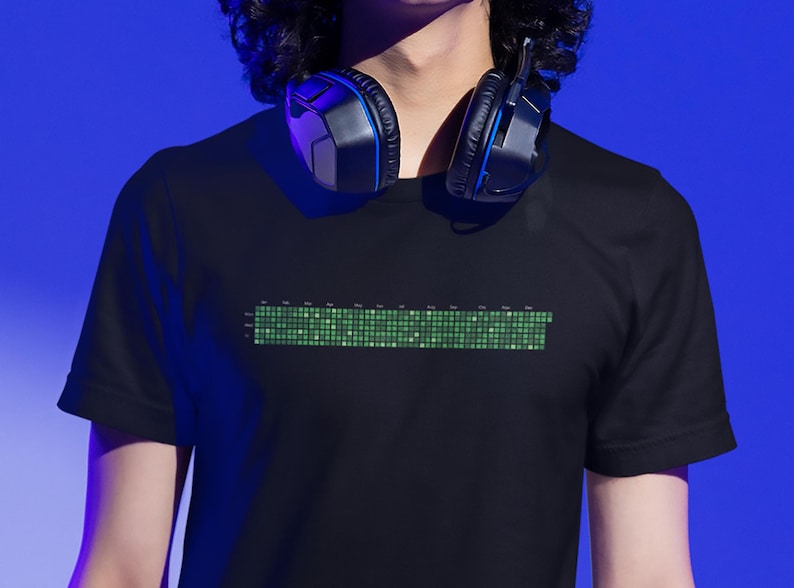 Może przedstawiać: Czarny T-shirt z zielonym pikselowym wzorem z przodu. Wz&oacute;r przypomina cyfrowy korektor dźwięku lub pasek postępu. Koszulkę nosi osoba z czarnymi włosami i słuchawkami na szyi.