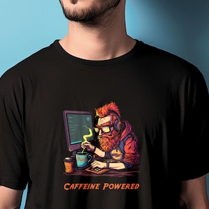 Op de afbeelding: Zwart T-shirt met een afbeelding van een roodharige man met een koptelefoon die koffie drinkt terwijl hij codeert op een computer. De tekst "CAFFEINE POWERED" staat onder de afbeelding.