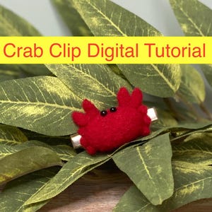 May include: A red felt crab clip with black eyes. The crab is attached to a silver hair clip. The crab is sitting on a bed of green leaves.