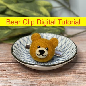 May include: A brown felt bear head attached to a silver hair clip. The bear has black eyes and a black nose. The clip is sitting on a white and blue patterned dish.