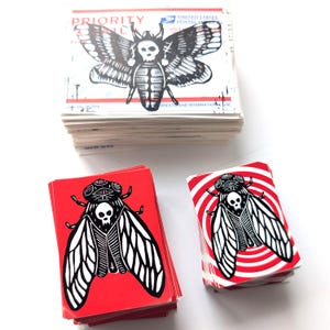 May include: A stack of red and white stickers with a black and white illustration of a cicada with a skull on its head. The cicada is on a red background with white concentric circles. The stickers are stacked on a white surface.