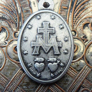 May include: Silver oval pendant with a cross and the letters 'A' and 'M' with two hearts below. The pendant has a textured surface and a loop at the top for hanging.