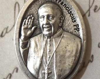 Może przedstawiać: Owalny, srebrzysty wisiorek z szczegółowym reliefem mężczyzny w stroju religijnym, podnoszącego prawą dłoń. Wisiorek jest ozdobiony napisem "Franciscus PP." i posiada małą pętlę na łańcuszek. Tło jest zapisane ręcznie.