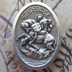May include: A silver oval pendant featuring St. George on horseback slaying a dragon. The pendant has the text "ST. GEORGE" at the top and a small loop for a chain. The background is a vintage paper with stamps and handwriting.