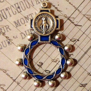 May include: A silver-toned metal pendant featuring a circular frame with blue enamel accents and small spherical beads along the outer edge. At the top, a cross-shaped element displays a central circular medallion with the text REGINA SINE LABE ORIGINALI CONCEPTA O.P.N. and a figure of the Virgin Mary.