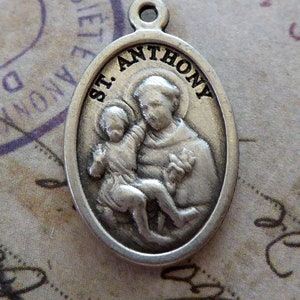 Puede incluir: Pendiente ovalado de plata con una representación de San Antonio sosteniendo un bebé. El pendiente tiene las palabras "ST. ANTHONY" inscritas alrededor del borde.