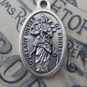 May include: Silver oval charm with a depiction of Our Lady Untier of Knots. The charm has a loop at the top for attaching to a necklace or bracelet.
