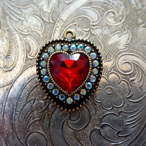 May include: A heart-shaped pendant featuring a large, faceted red center stone surrounded by a border of small, iridescent clear crystals set in an antique bronze-toned metal frame with a small circular loop at the top for attachment.