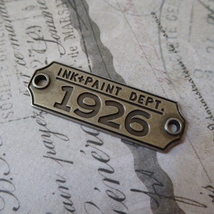 May include: A silver-toned metal tag with rounded corners and two holes. The tag has the text "INK+PAINT DEPT." and the year "1926" embossed on it.