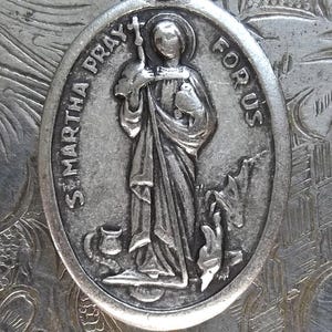 Pu&ograve; includere: Ciondolo ovale in argento con immagine incisa di Santa Marta che tiene una croce e un oggetto, con la scritta "ST. MARTHA PRAY FOR US". Il ciondolo ha un anello nella parte superiore per appenderlo. Sfondo floreale.