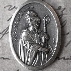 May include: An oval-shaped silver-toned metal pendant featuring a relief of Saint Patrick wearing a miter and holding a crozier. The text "ST. PATRICK" is embossed on the left side, and the word "ITALY" is stamped on the right side of the medal.