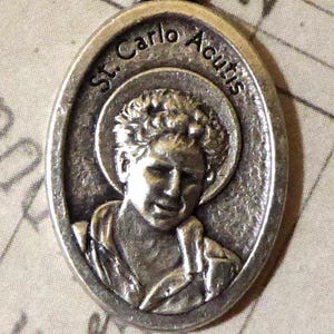 Puede incluir: Un colgante ovalado de metal plateado con un retrato en relieve de una persona joven con cabello rizado y un halo. El texto St. Carlo Acutis está grabado a lo largo del borde superior curvo del colgante, que incluye una anilla en la parte superior.