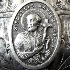 May include: Oval silver pendant featuring St. Francis Xavier holding a crucifix, with the inscription "St. Francis Xavier P.F.U." around the image. The pendant has a loop at the top for hanging and is set against a textured background.