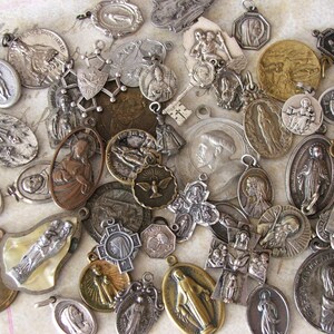 May include: A collection of vintage silver and gold religious medallions featuring various images of saints, crosses, and religious symbols. The medallions are in various shapes and sizes and have a worn, antique look.