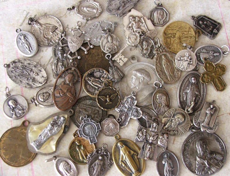 May include: A collection of vintage silver and gold religious medallions featuring various images of saints, crosses, and religious symbols. The medallions are in various shapes and sizes and have a worn, antique look.