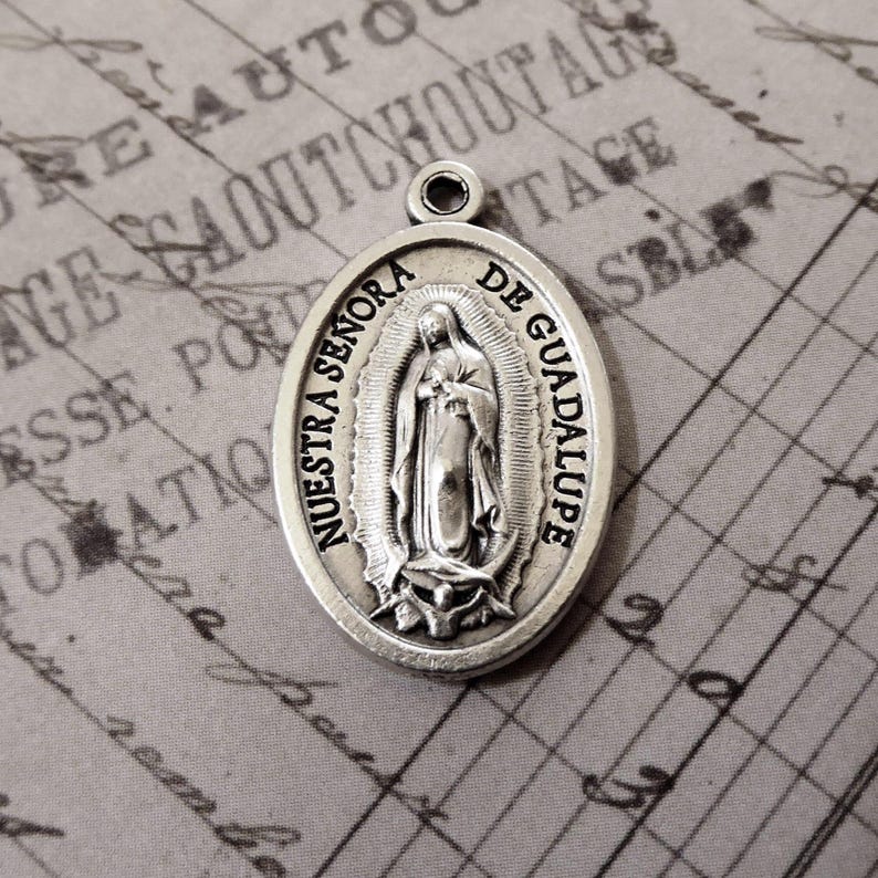 K&ouml;nnte beinhalten: Ovaler Silberanh&auml;nger mit dem Bild Unserer Lieben Frau von Guadalupe, mit dem Text "Nuestra Se&ntilde;ora de Guadalupe". Der Anh&auml;nger hat eine kleine Schlaufe f&uuml;r eine Kette. Der Hintergrund ist ein Vintage-Papier.