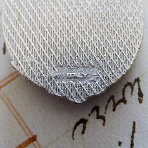 May include: Close-up of the back of a silver charm with a textured surface and the word "ITALY" stamped on the bottom.