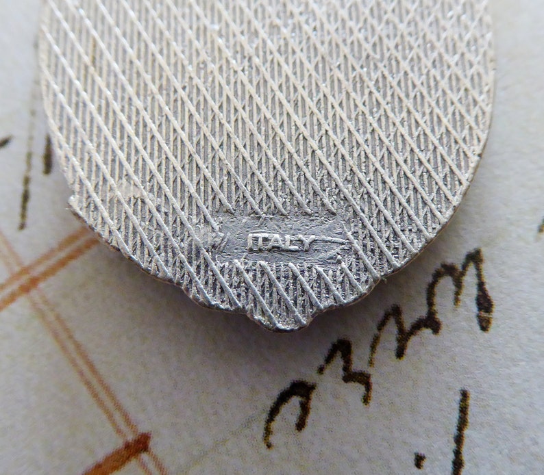 May include: Close-up of the back of a silver charm with a textured surface and the word "ITALY" stamped on the bottom.