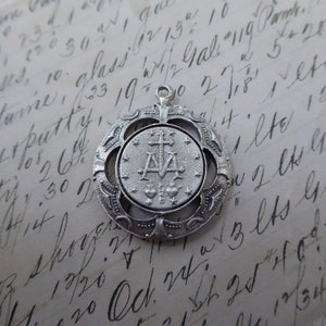 May include: Silver pendant with a detailed design featuring a cross and the letters 'A' and 'M' surrounded by stars. The pendant is on a white background with handwritten text.