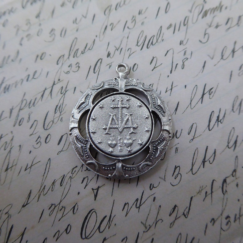 May include: Silver pendant with a detailed design featuring a cross and the letters 'A' and 'M' surrounded by stars. The pendant is on a white background with handwritten text.