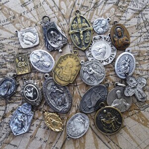 K&ouml;nnte beinhalten: Eine Sammlung religi&ouml;ser Anh&auml;nger und Medaillons in verschiedenen Formen und Gr&ouml;&szlig;en. Die Anh&auml;nger sind aus silber- und goldfarbenem Metall gefertigt und zeigen detaillierte religi&ouml;se Figuren und Symbole. Der Hintergrund ist eine Vintage-Karte.