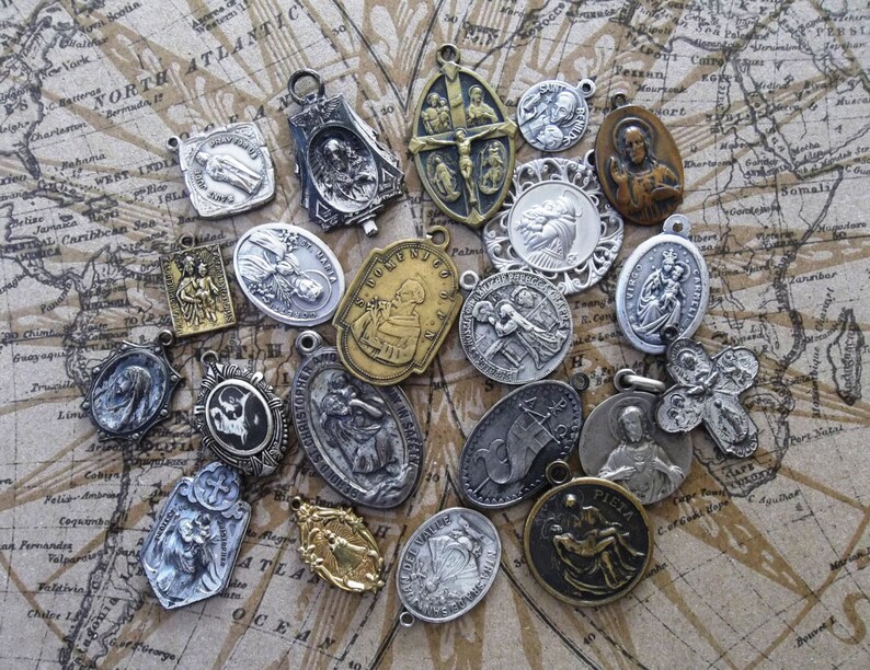 K&ouml;nnte beinhalten: Eine Sammlung religi&ouml;ser Anh&auml;nger und Medaillons in verschiedenen Formen und Gr&ouml;&szlig;en. Die Anh&auml;nger sind aus silber- und goldfarbenem Metall gefertigt und zeigen detaillierte religi&ouml;se Figuren und Symbole. Der Hintergrund ist eine Vintage-Karte.