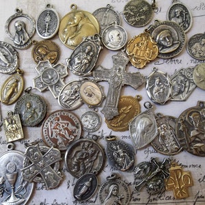 May include: A collection of vintage religious medallions, charms, and pendants. The medallions are made of metal and feature various religious symbols, including crosses, saints, and the Virgin Mary. Some of the medallions are gold-toned, while others are silver-toned.