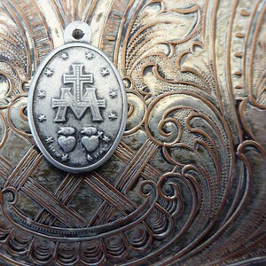 May include: Silver-toned metal pendant with a religious design. The pendant features a cross with the letters "M" and "A" on either side. Two hearts are below the cross. The pendant is on a textured silver background.