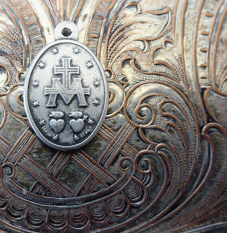 May include: Silver-toned metal pendant with a religious design. The pendant features a cross with the letters "M" and "A" on either side. Two hearts are below the cross. The pendant is on a textured silver background.