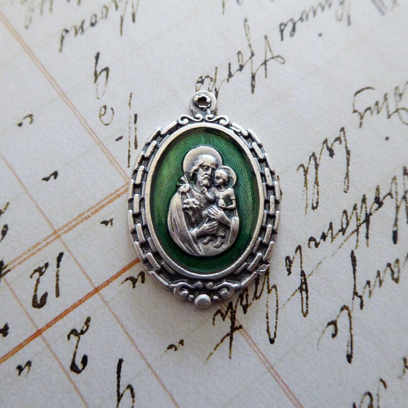 May include: Silver oval pendant with a green enamel background depicting a religious figure holding a child. The figure is wearing a robe and has a halo. The child is also wearing a robe and has a halo.