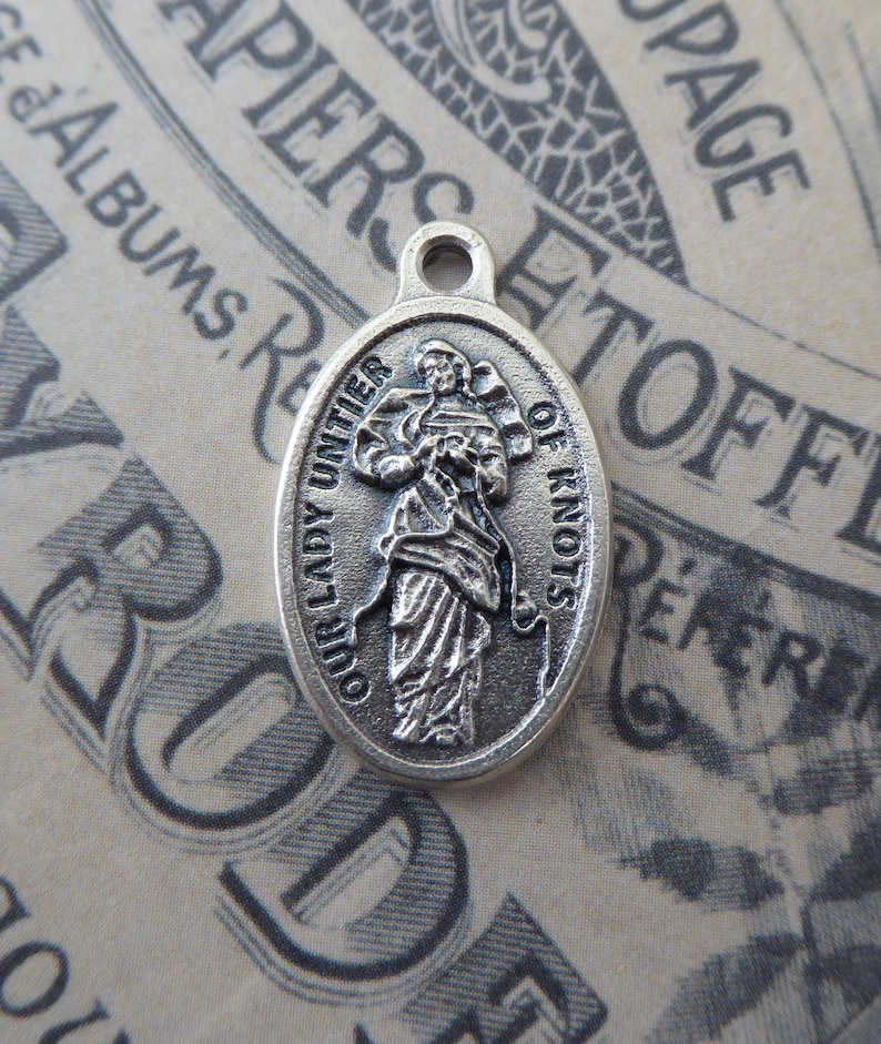 May include: Silver oval charm with a depiction of Our Lady Untier of Knots. The charm has a loop at the top for attaching to a necklace or bracelet.