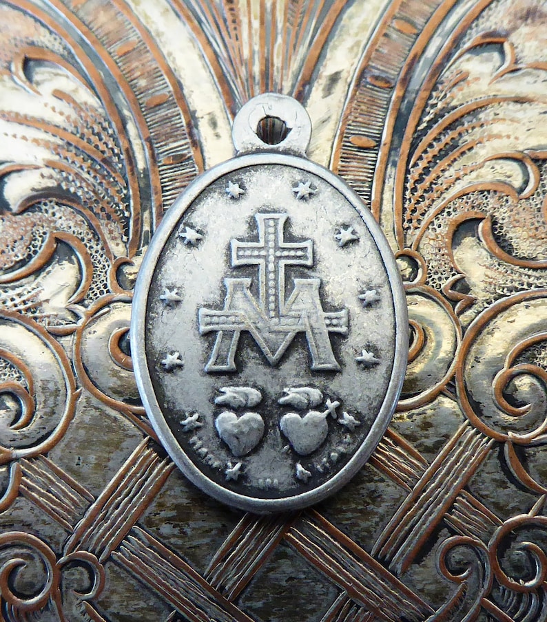 May include: Silver oval pendant with a cross and the letters 'A' and 'M' with two hearts below. The pendant has a textured surface and a loop at the top for hanging.