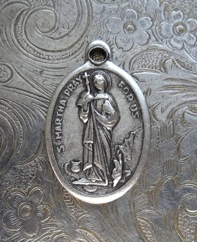 Pu&ograve; includere: Ciondolo ovale in argento con immagine incisa di Santa Marta che tiene una croce e un oggetto, con la scritta "ST. MARTHA PRAY FOR US". Il ciondolo ha un anello nella parte superiore per appenderlo. Sfondo floreale.