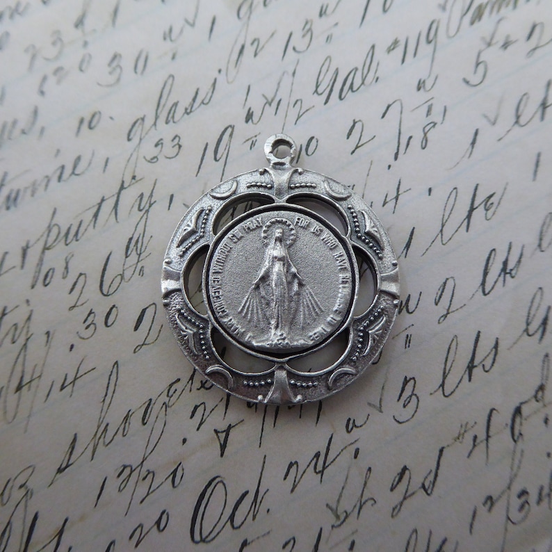 May include: Silver pendant featuring a detailed image of the Virgin Mary with the text "RECEIVED WITHOUT SPRAY FOR US WHO HAVE RECOURSE TO THEE" around the image.