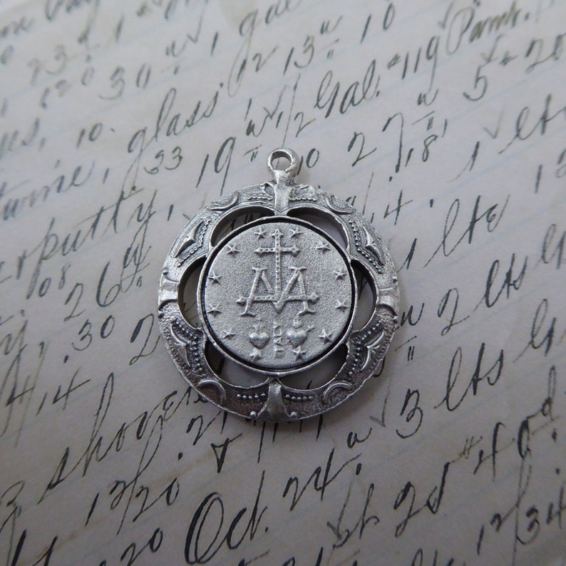 May include: Silver pendant with a detailed design featuring a cross and the letters "M.A." surrounded by stars. The pendant is attached to a silver chain.