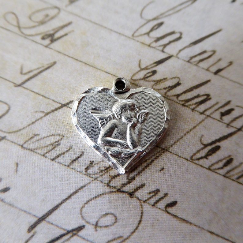 May include: Silver heart-shaped charm with an angel design. The angel is depicted with wings and holding a small object. The charm has a textured surface.