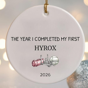 May include: A round white ceramic ornament hanging by a gold string. The text reads THE YEAR I COMPLETED MY FIRST HYROX, followed by a small illustration of a sled and a medicine ball, and the year 2026 printed at the bottom of the ornament.